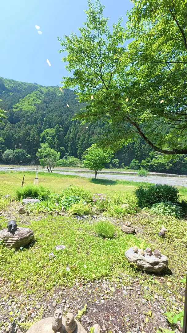 窓の外。焼き物の動物が置かれた庭の先に草地と河原、その向こうに山